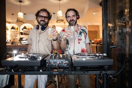 Deux DJs souriants tenant des verres, avec des platines et une ambiance festive en arrière-plan.